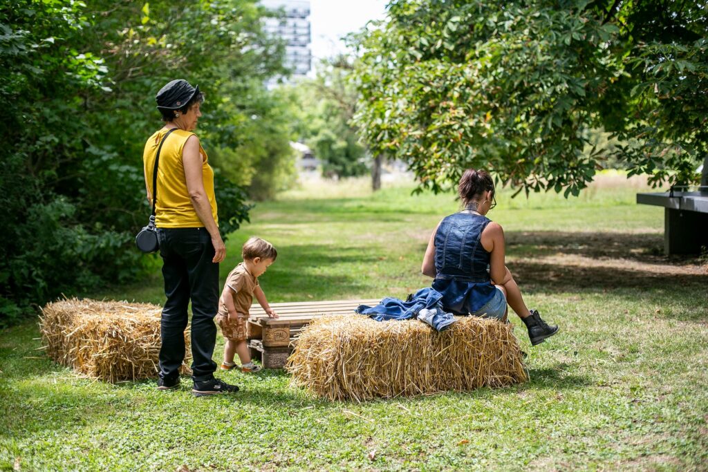 Une femme est assise sur une botte de paille, à côté d'elle se trouvent un petit enfant et une dame âgée vêtue d'un t-shirt jaune.