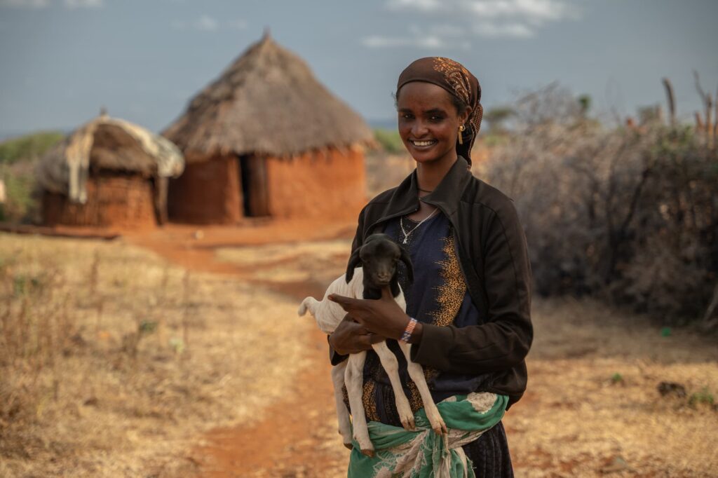 Une femme tenant une petite chèvre dans ses bras devant une hutte.